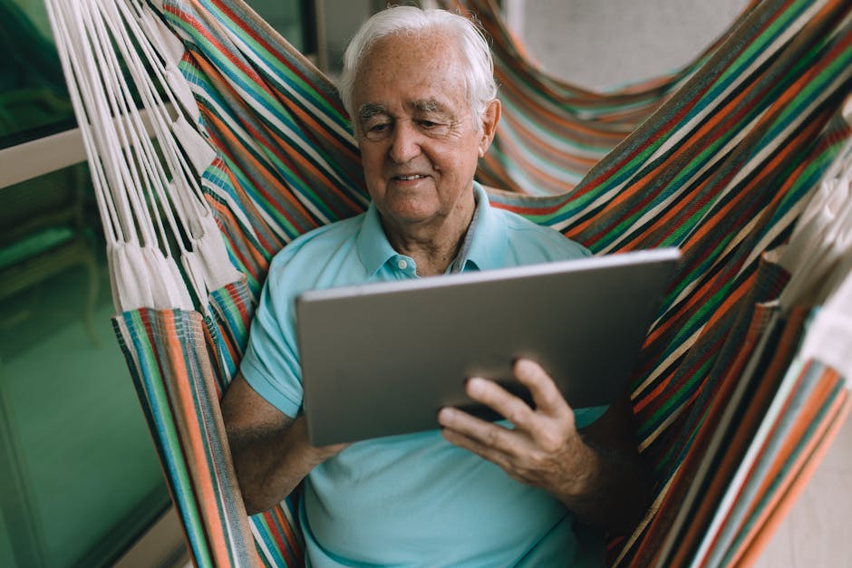 Elderly man enjoying leisure time in hammock while using a tablet indoors.