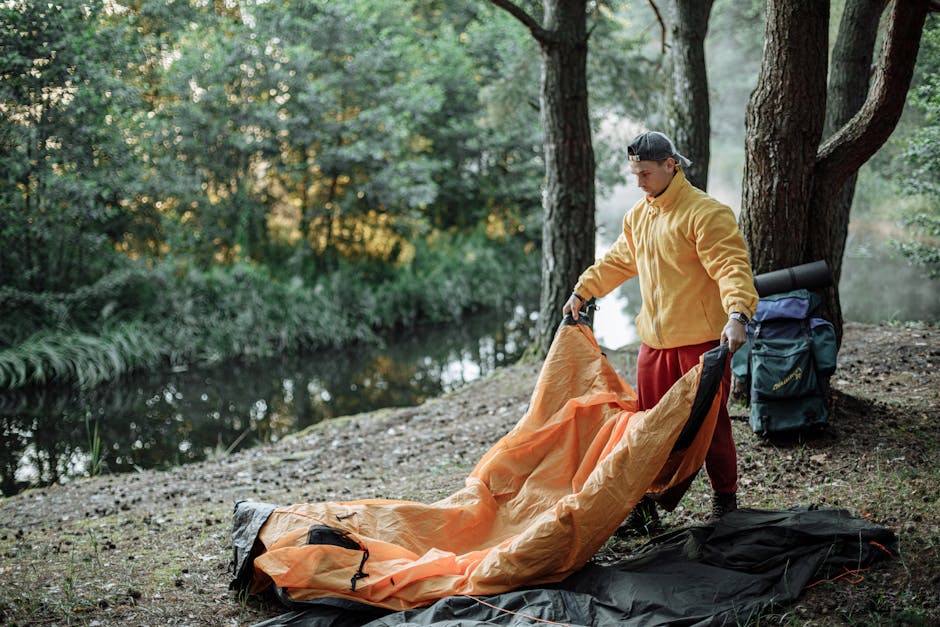 A man sets up an orange tent near a river in the forest, capturing the essence of outdoor adventure.