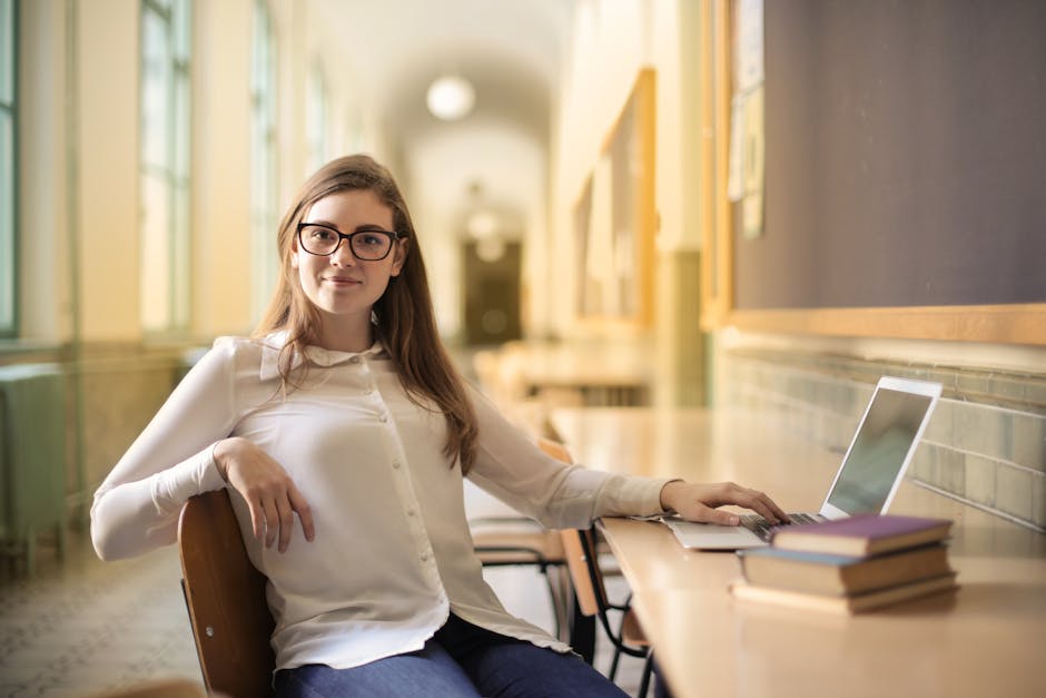 Smiling young woman using a laptop in a study area, wearing glasses and white blouse.