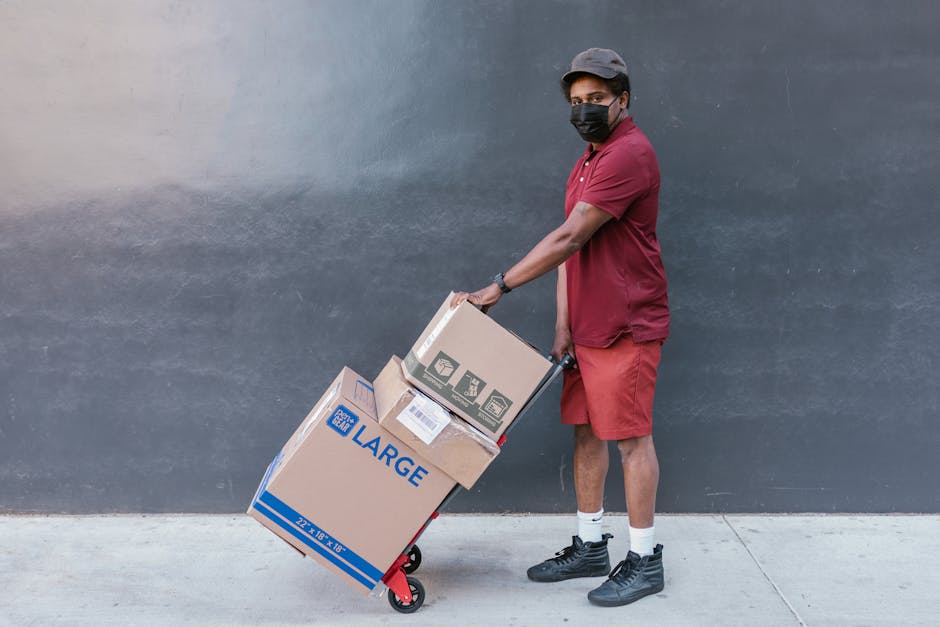 Delivery worker wearing face mask transporting boxes on a cart outside. Safe and essential service during COVID-19 pandemic.