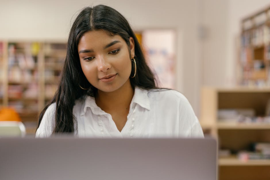 A thoughtful woman using a laptop in a library setting, focused on her work.