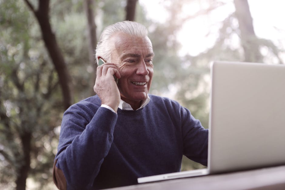 Elderly man smiling while on phone call with laptop outdoors.