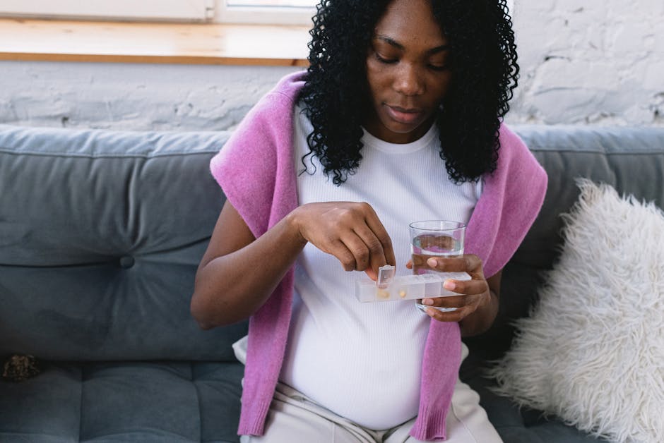 Serious pregnant African American female with glass of water taking prenatal vitamins while sitting on comfortable sofa in living room