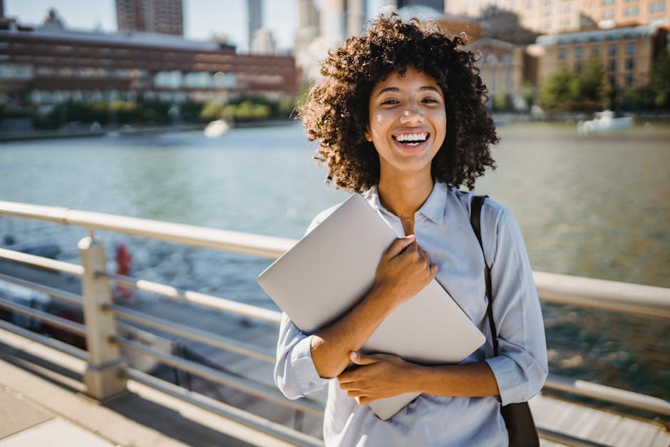 Smiling young woman with curly hair holding a laptop by the river on a sunny day.