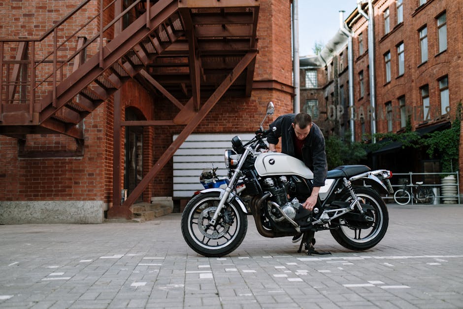 A man repairs a white motorcycle outside brick buildings, highlighting urban life and motorcycle maintenance.