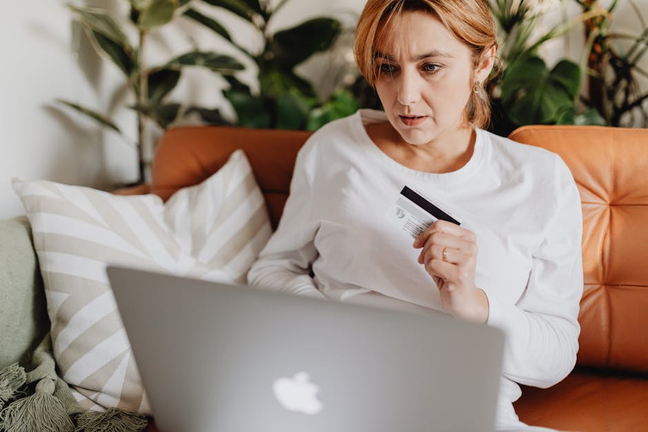 A woman sitting on a couch shopping online with a credit card and laptop in a cozy living room.