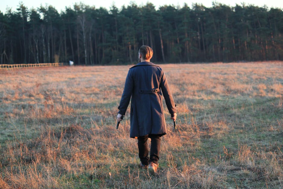 A man in a long coat walking through a grassy field holding guns, forest in background.