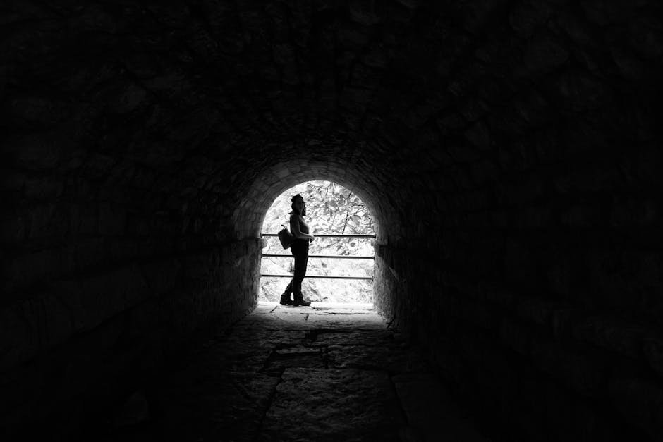 Silhouette of a person standing in a stone tunnel, with light at the end and a rustic atmosphere.