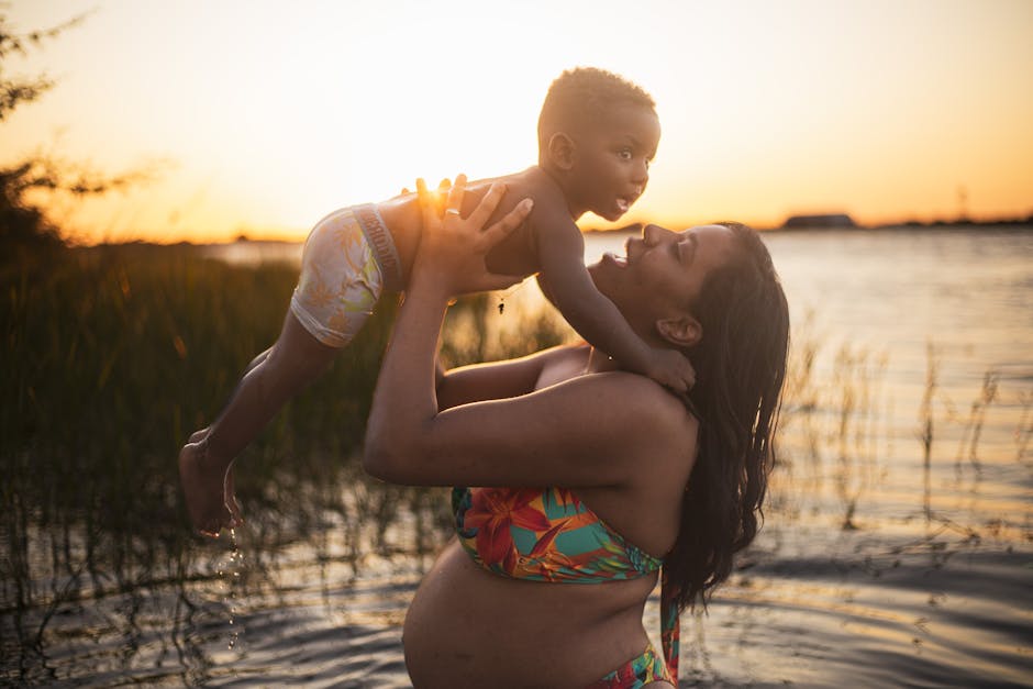 Pregnant woman holds baby by the water at sunset, showcasing family love and joy.
