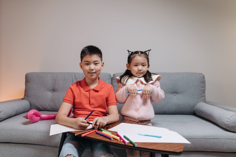 Asian siblings smile while drawing with colorful markers in a cozy indoor setting.