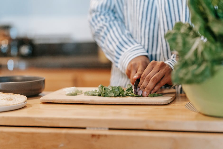 Crop anonymous ethnic male in striped shirt cutting fresh spinach on chopping board while cooking at home