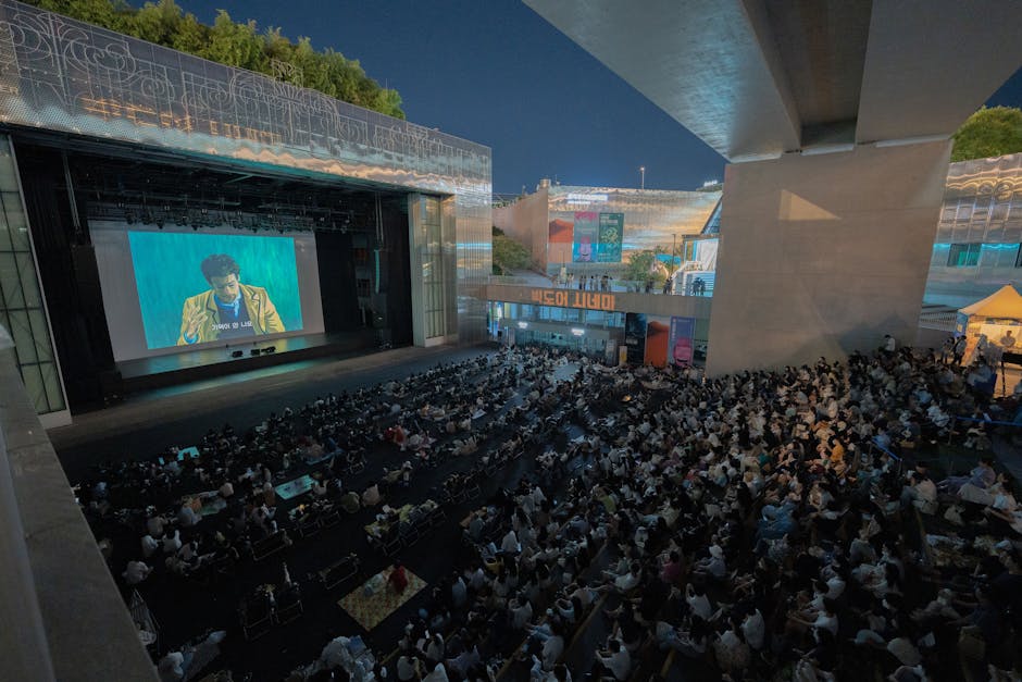 A large audience gathers outdoors at night to watch a movie screening under a bridge.