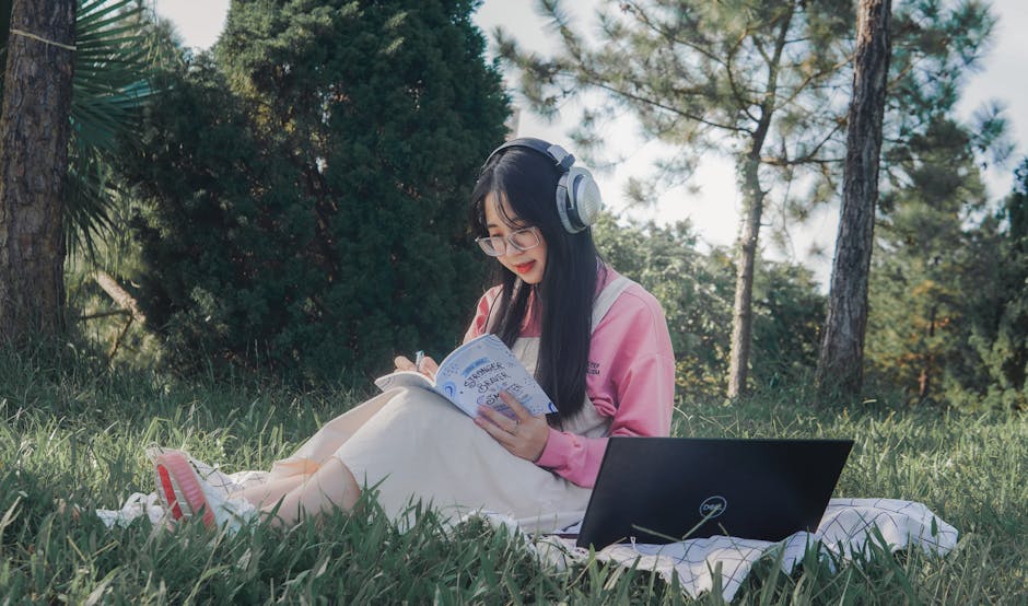 Asian woman studying outdoors with headphones, notebook, and laptop on the grass under trees.