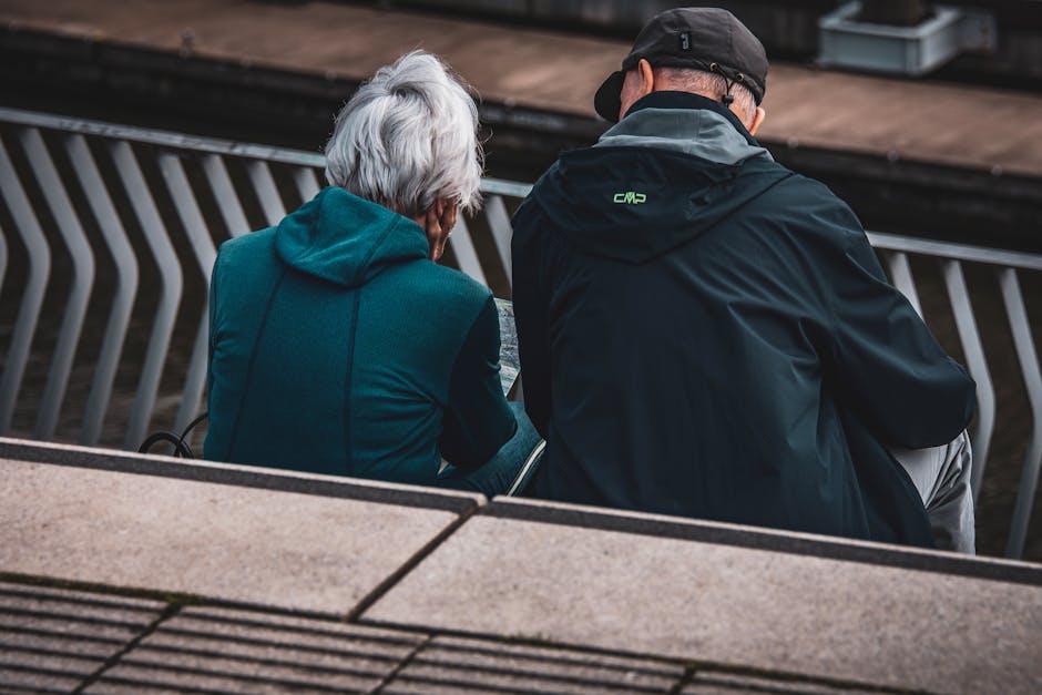 Elderly couple in jackets sitting on steps overlooking an urban setting, captured candidly from behind.