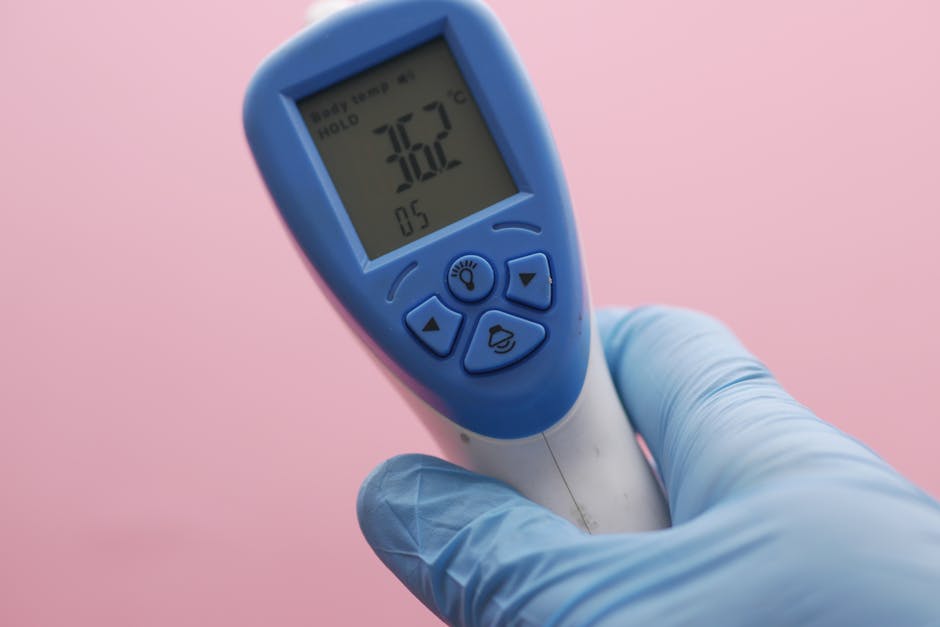 Close-up of an infrared thermometer displaying temperature, held by a gloved hand against a pink background.