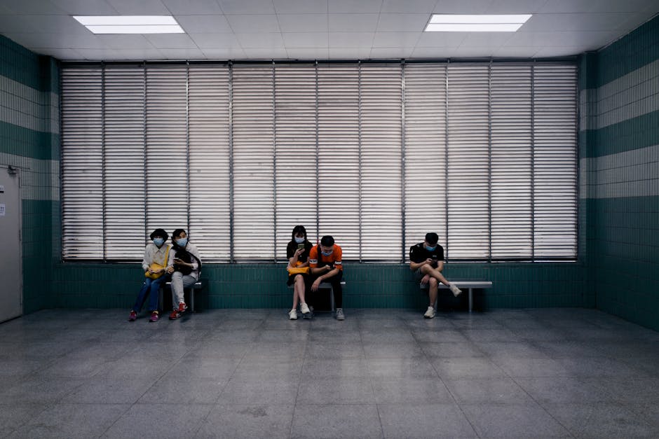 Full body of unrecognizable people in medical masks sitting on benches in spacious hall of clinic against windows