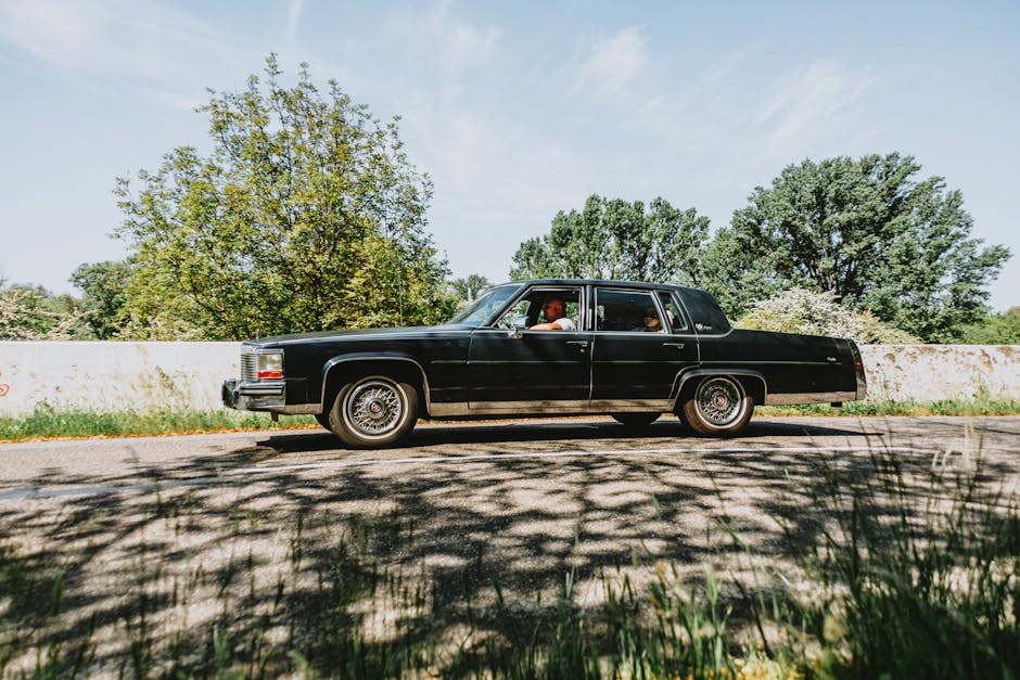 Vintage black sedan cruising on a scenic road on a sunny day, surrounded by lush greenery.