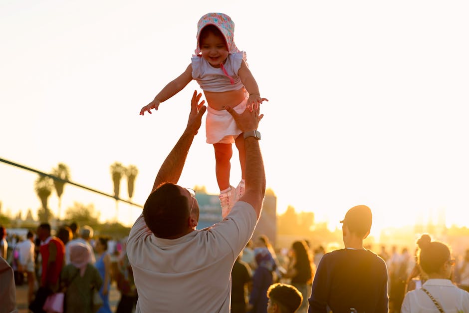 A joyful moment as a father lifts his daughter into the air at a sunny outdoor festival.