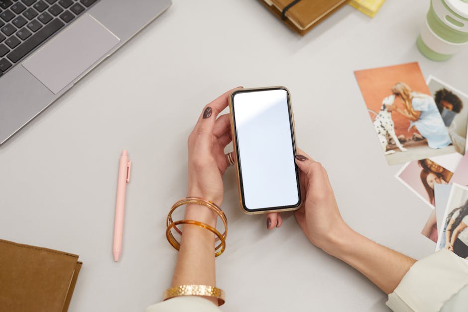 Close-up of a woman holding a smartphone on a desk with a laptop and photos, capturing modern workspace vibes.