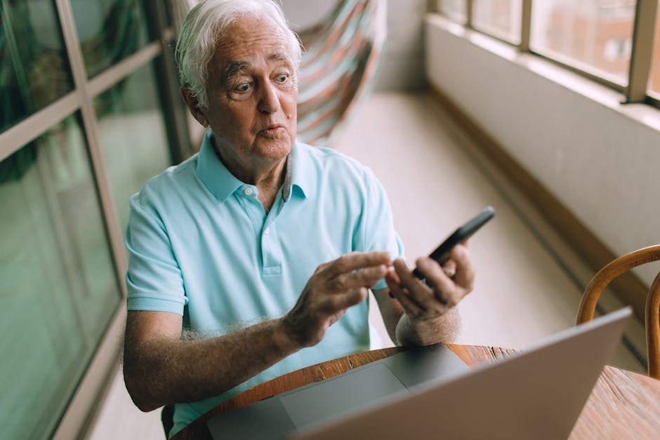 Senior man sitting indoors, using smartphone and laptop, emphasizing technology connection.