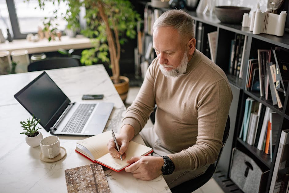 A mature man with a beard writes in a diary at his home office desk next to a laptop.