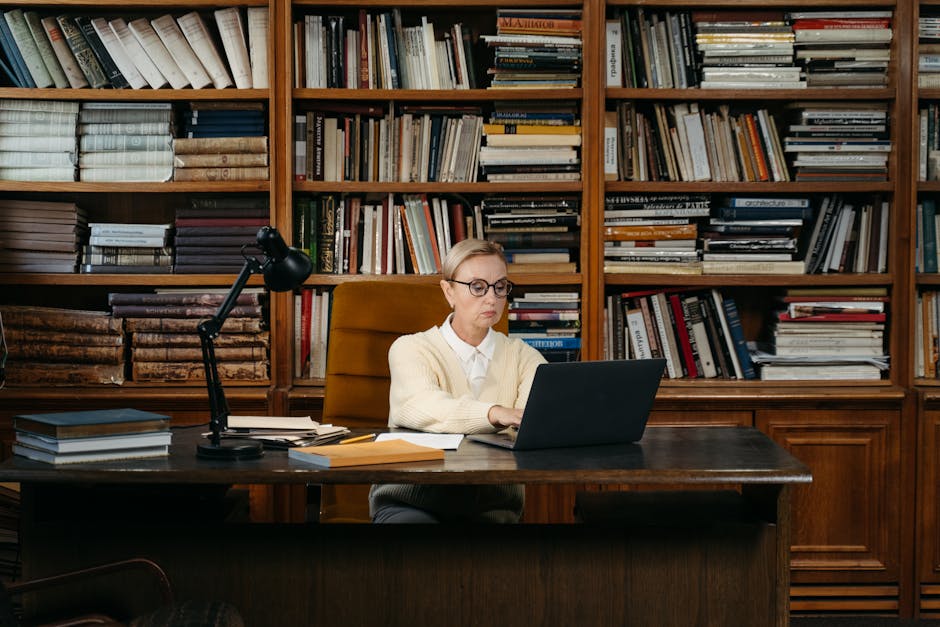 Woman in a yellow sweater working on a laptop amidst books in a library setting.