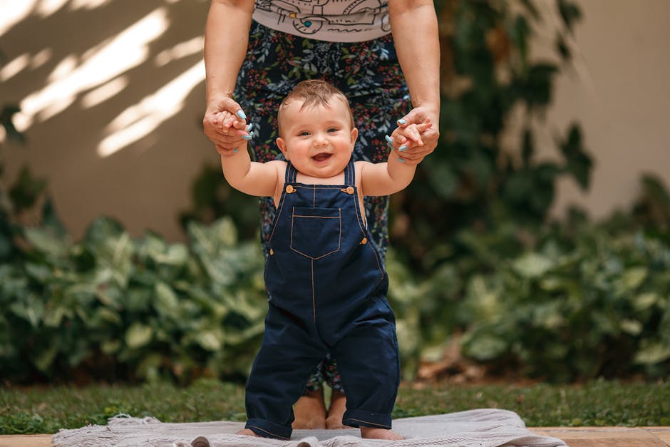 Cheerful baby in overalls being supported while taking first steps outdoors.