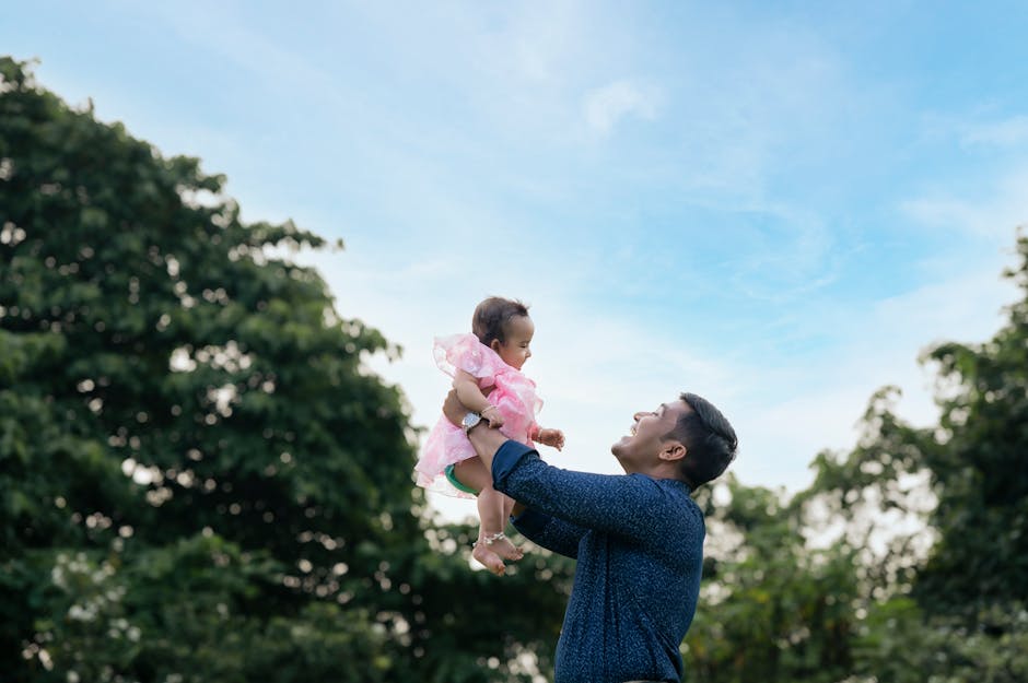 A joyful moment of a father lifting his baby girl outdoors against a clear blue sky.