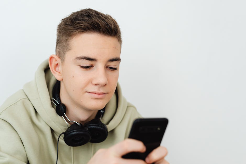 Young man in a hoodie using a smartphone with headphones around his neck in a bright setting.