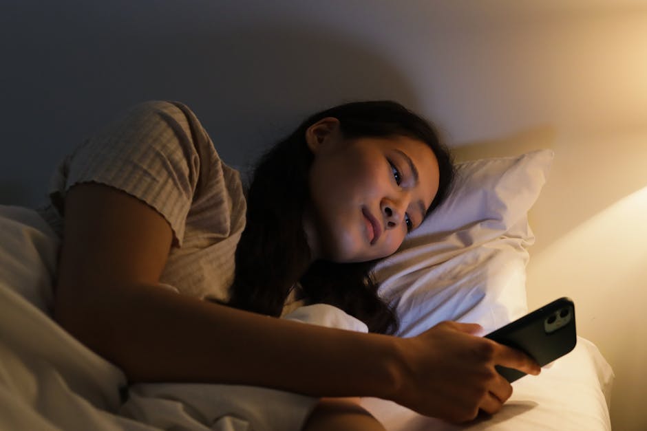 An adult woman lying in bed using a smartphone, illuminated by warm bedside lighting.