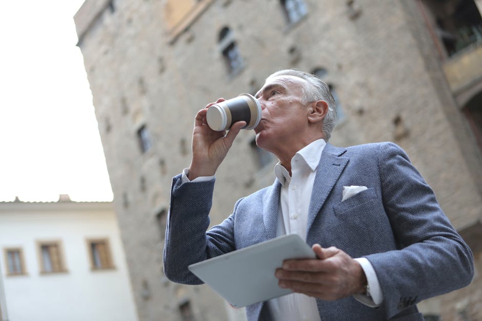 Elderly man in a suit drinks coffee outdoors while holding a tablet.