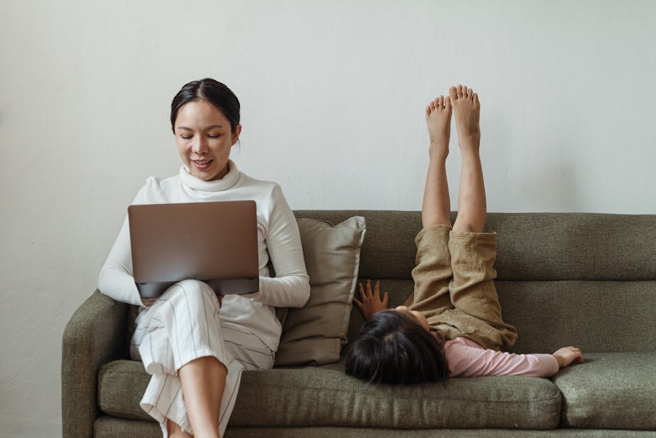Positive smiling Asian woman in casual clothes sitting on sofa and browsing netbook near cute daughter lying with raised legs