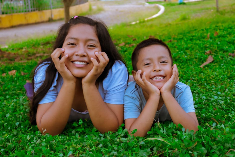A cheerful duo of a teenager and a child laying on grass, sharing joyful smiles outdoors.