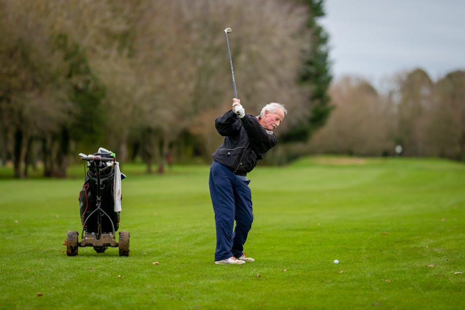 Elderly man swinging a golf club on a vibrant golf course with trolley in background.