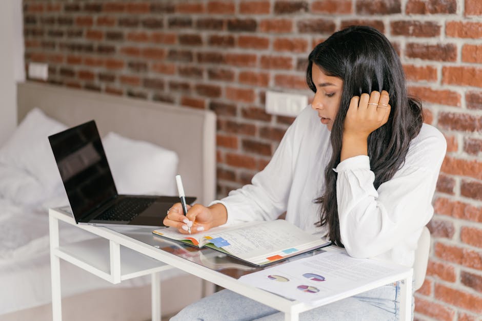 A young woman studies indoors with a laptop, writing notes in a notebook on a desk.
