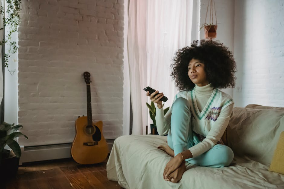 Full body of relaxed young African American female millennial with curly hair in casual clothes sitting on couch and switching TV channels with remote controller during weekend at home