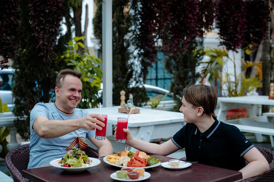 Father and son clink glasses over food and drinks outdoors, enjoying a summer meal together.