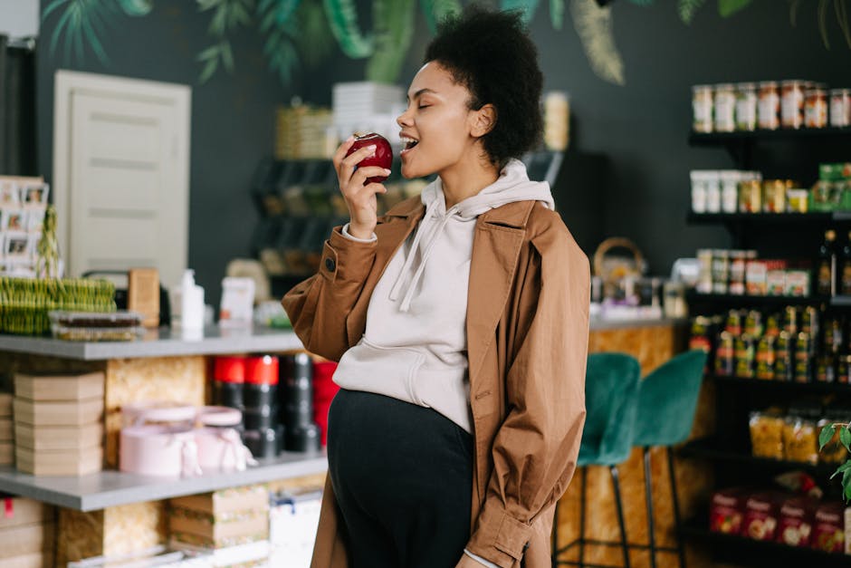 Pregnant woman with afro hair enjoying a fresh apple inside a grocery store.