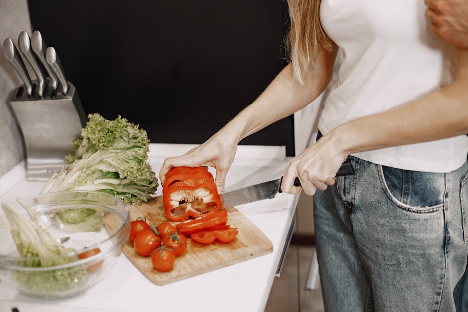 Close-up of a person slicing red bell pepper and tomatoes on a wooden chopping board indoors.