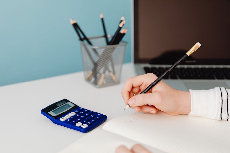 Close-up of a hand writing in a notebook beside a calculator and laptop on a desk.