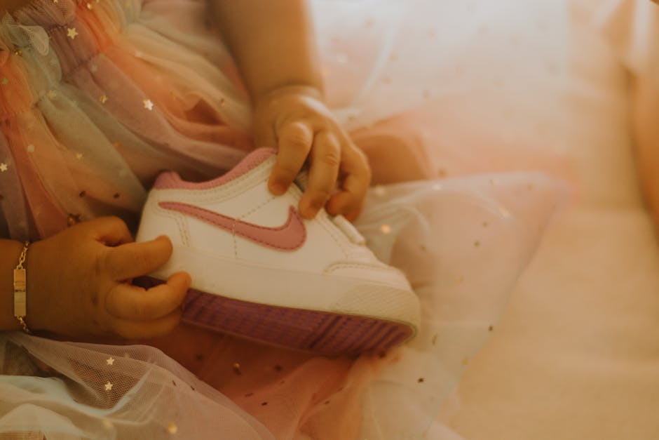 A baby in a multicolored fairy-tale dress holding a pink and white baby shoe, with a warm and soft tone.