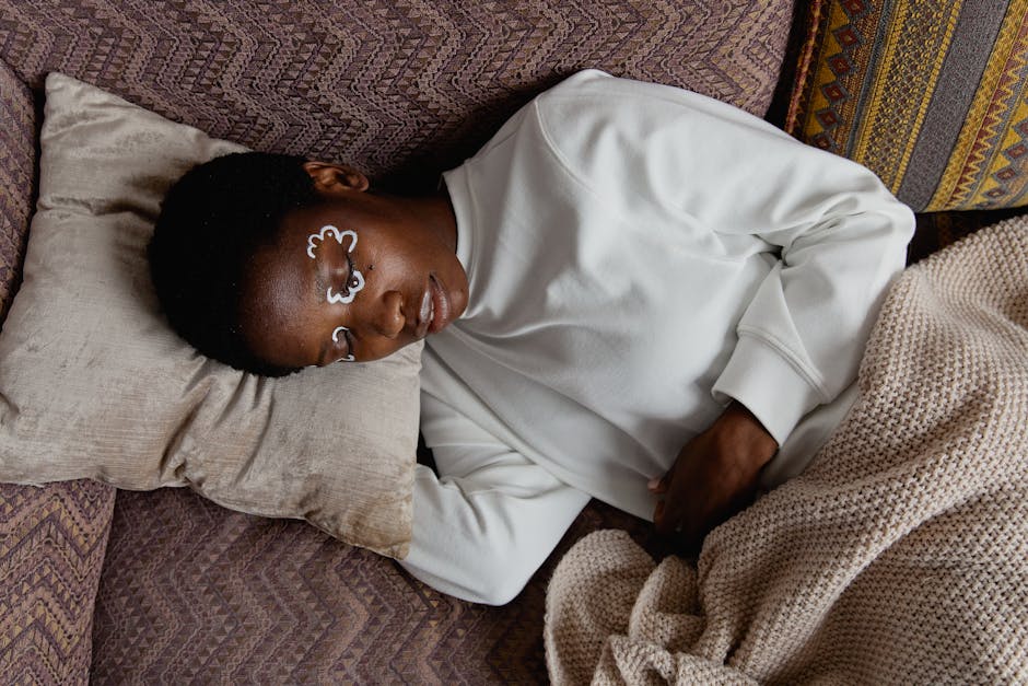 An adult woman relaxing indoors with artistic face paint, lying on a pillow and blanket in a cozy setting.