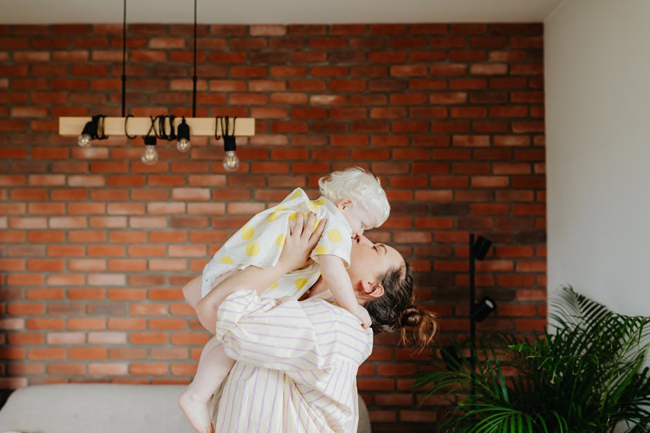A mother lovingly lifts her child against a textured brick wall indoors.