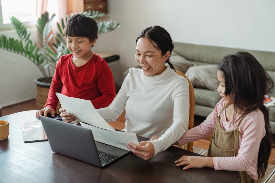 From above of smiling happy Asian mother sitting at table with laptop and watching pictures painted by adorable little son and daughter