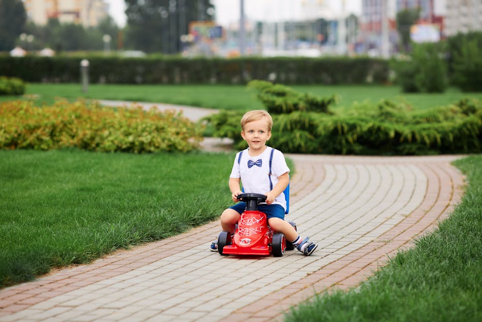 A smiling child rides a red toy car on a brick path in a green park, enjoying a fun day outdoors.