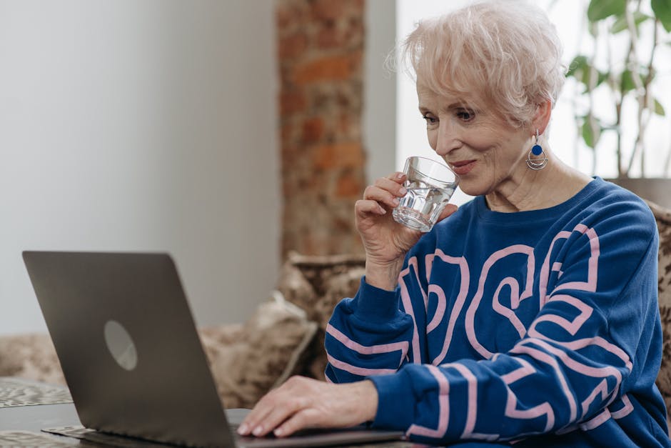 Elderly woman sipping water while working on a laptop in a cozy indoor setting.