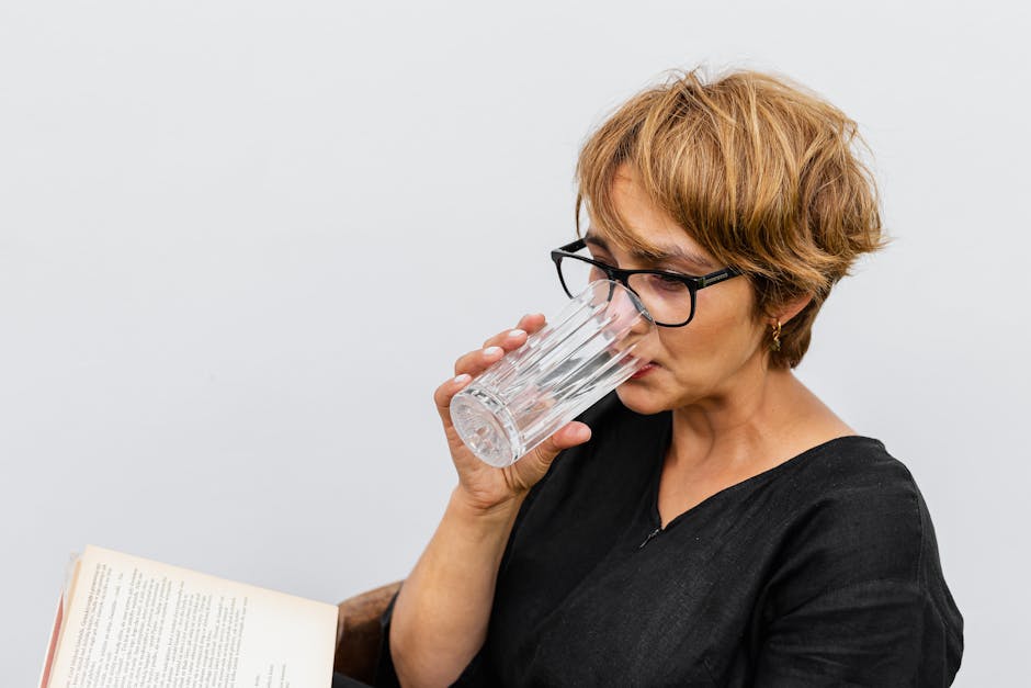 A woman with eyeglasses reading a book and drinking water indoors.
