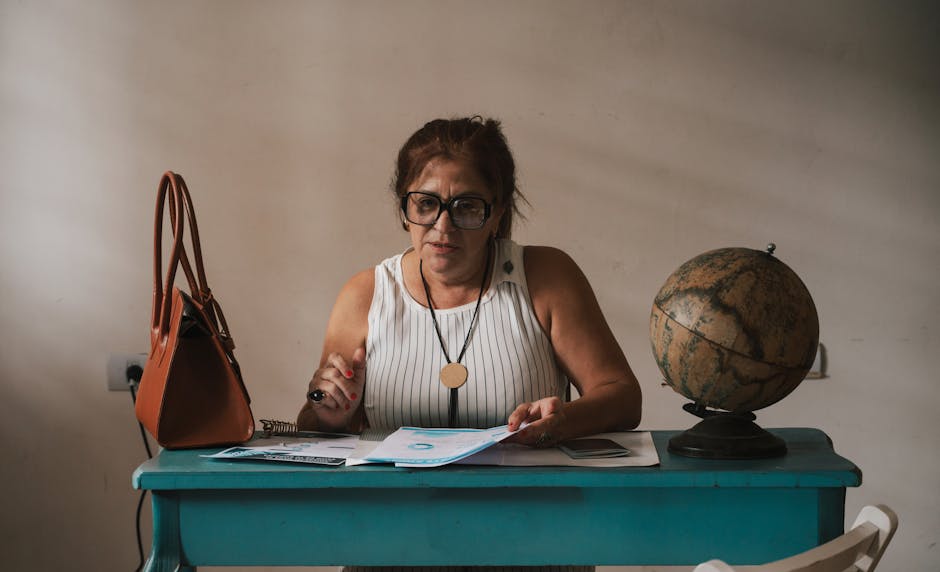 A middle-aged woman sits at her home desk with documents, a globe, and a bag.
