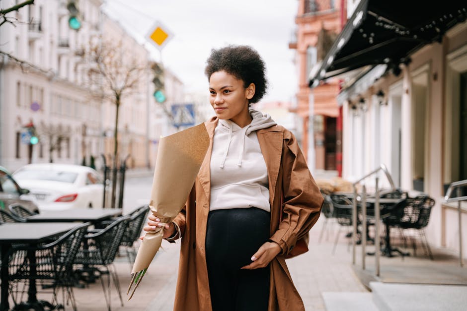 A stylish pregnant woman with afro hair holding flowers, walking down a charming city street.