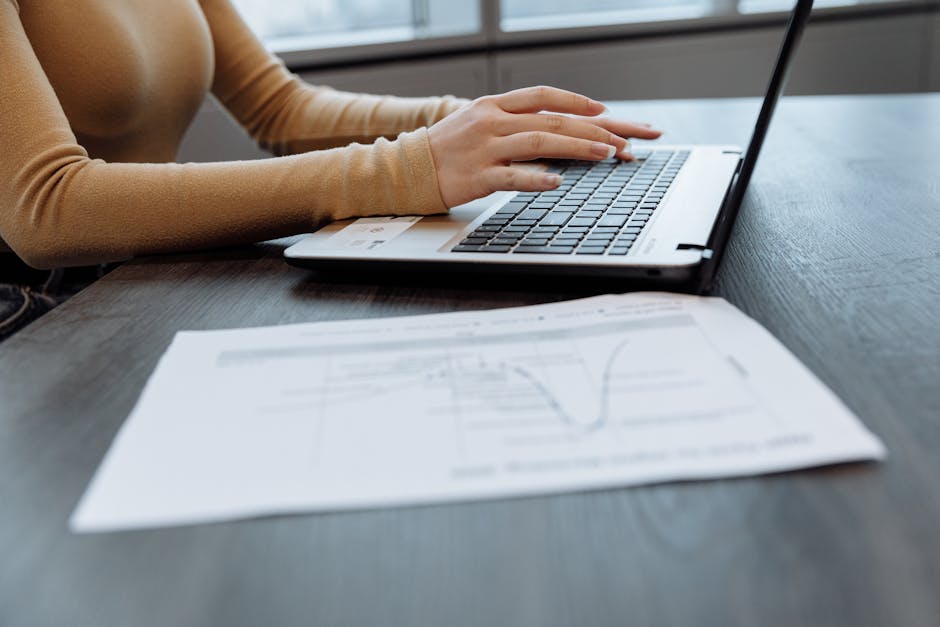 Close-up of a person's hands typing on a laptop at a desk with documents. Ideal for office, work, and technology themes.